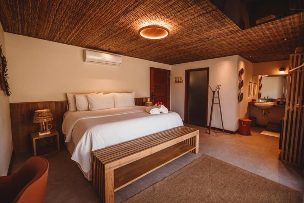 Um quarto da Pousada Moana aconchegante com uma cama grande, móveis de madeira, teto de vime e um banheiro visível através de uma porta aberta. Imagem para ilustrar post sobre onde ficar em Fernando de Noronha.