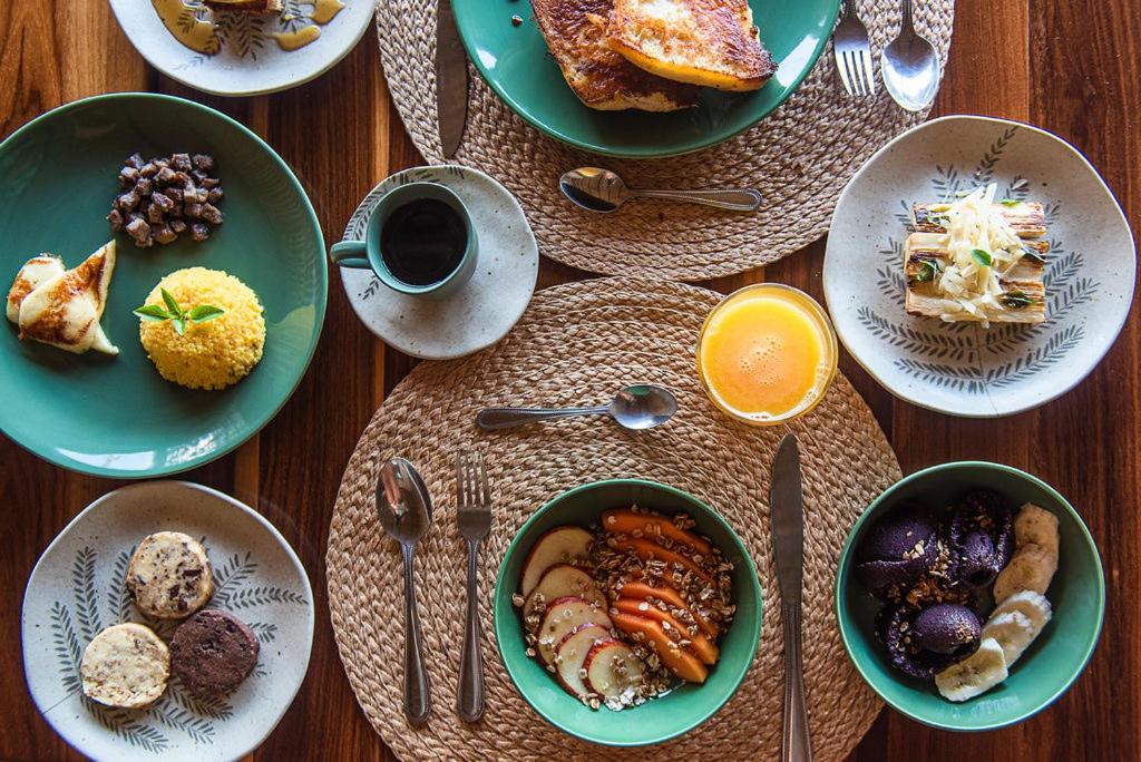 Uma mesa de madeira da Pousada Sela Gineta com pratos com alimentos diversos, incluindo frutas, bolo, pudim e uma bebida, dispostos em jogos americanos de tecido.