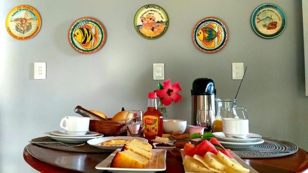 Uma mesa de jantar com doces, café, suco, frutas e xarope na Pousada Ventos da Atalaia. Pratos decorativos de peixe estão na parede cinza atrás da mesa. Imagem para ilustrar post sobre onde ficar em Fernando de Noronha.