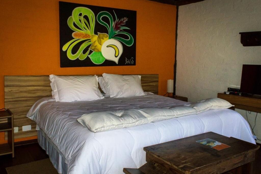 Quarto da Pousada Vila Santa Barbara. Uma cama de casal está no meio, em frente um móvel de madeira. Ao seu lado direito é possível ver a tv.