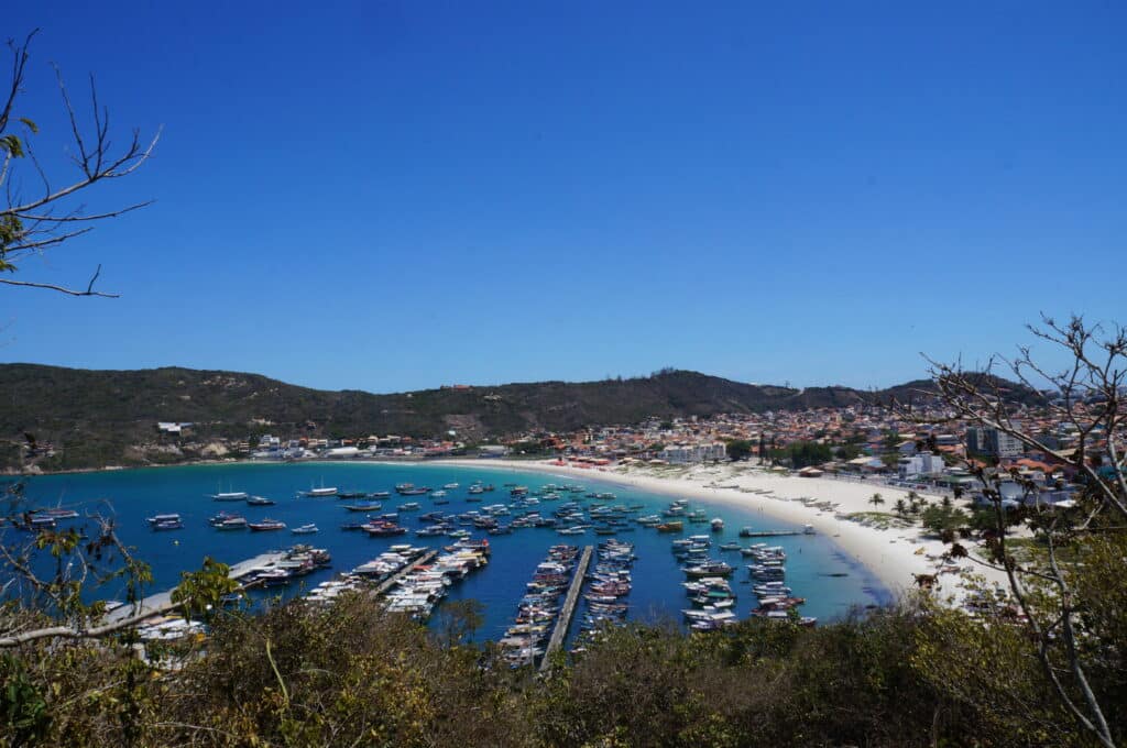 A foto mostra a Praia dos Anjos. Ela tem um porto movimentado cheio de barcos, uma praia de areia e colinas ao fundo, sob um céu azul claro.