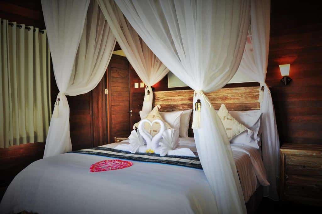 Um quarto aconchegante do Ocean Terrace Suite And Spa Luxurycom uma cama de dossel adornada com cortinas brancas. Cisnes feitos de toalhas e pétalas de flores decoram a cama. Representa onde ficar em Bali.