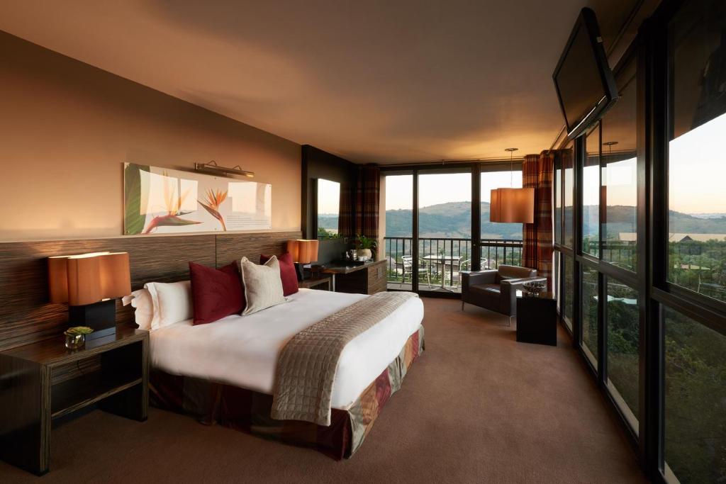 Um quarto do Thaba Eco Hotel moderno com uma cama queen-size, duas luminárias, uma escrivaninha com uma cadeira e grandes janelas com vista panorâmica. Representa Joanesburgo.