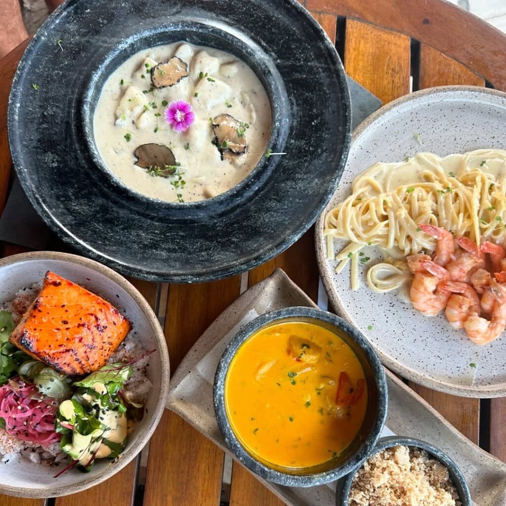 Mesa do Sol na cozinha, restaurante em Arraial do Cabo. A mesa carrega os pratos: macarrão cremoso com carne, camarão com macarrão, salmão com salada e uma tigela de curry amarelo.