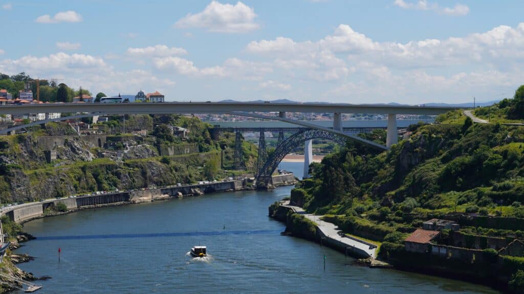 Rio Douro com um barco navegando, duas pontes, cercado por colinas verdes e edifícios, sob um céu parcialmente nublado. Representa um dos lugares do Roteiro para Portugal