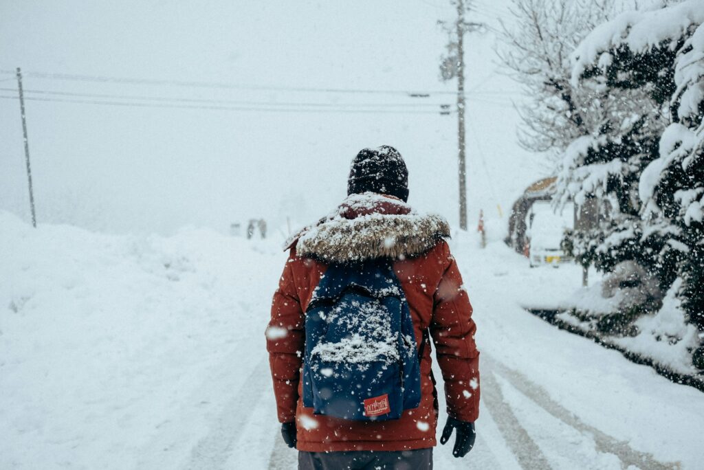 Pessoa vestindo um casaco vermelho e mochila caminhando em uma rua com neve, com árvores cobertas de neve e linhas de energia ao fundo em Shirakawago, uma vila no Japão.