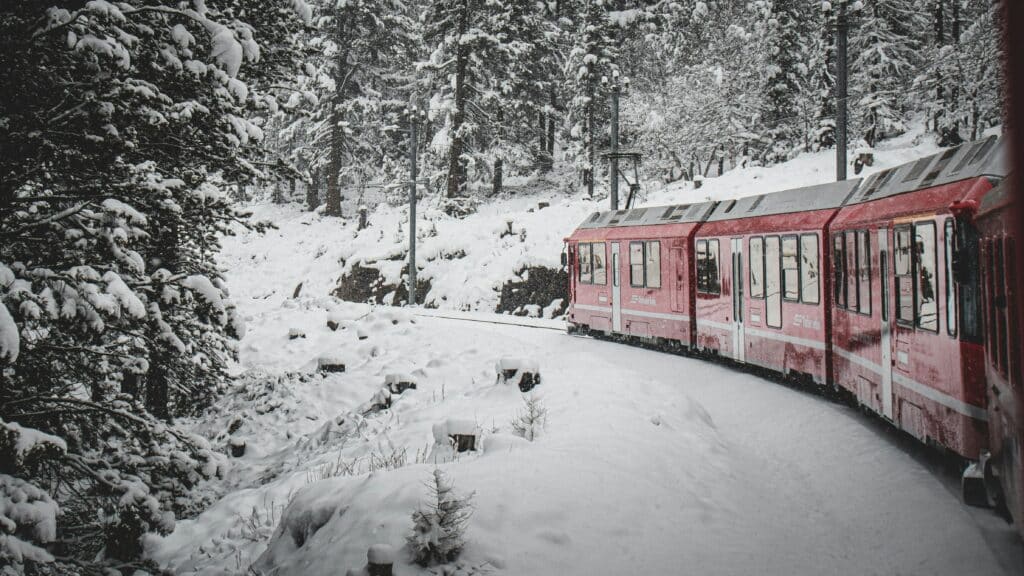 Um trem vermelho em St Moritz viaja por uma paisagem de floresta nevada, cercado por árvores cobertas de neve e um manto branco no chão. Representa como viajar de trem pela Suíça.