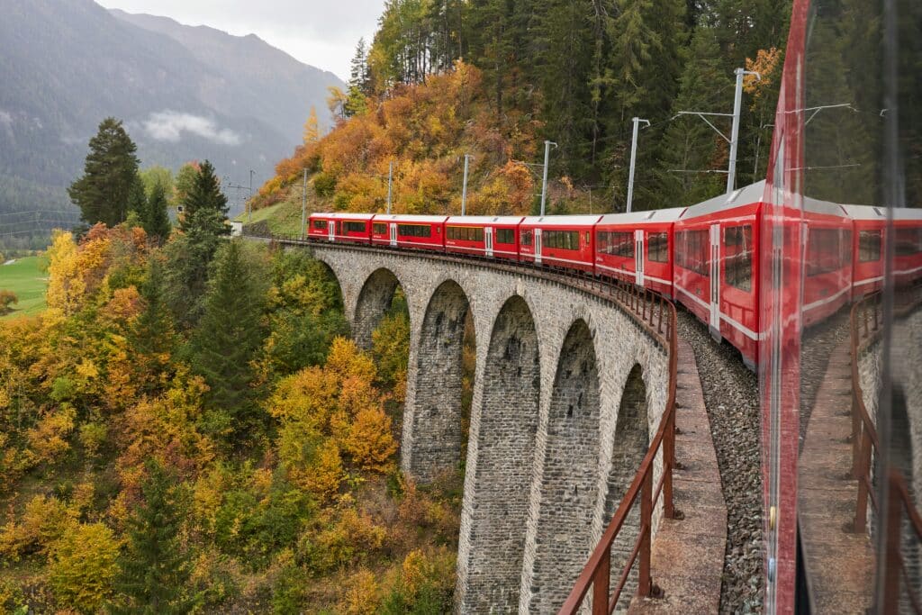 Um trem vermelho em Albula viaja por um viaduto de pedra curvo cercado por folhagens de outono com montanhas ao fundo. Representa como viajar de trem pela Suíça.