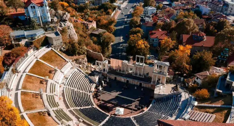 Vista aérea de um antigo anfiteatro romano em uma cidade, cercado por árvores e edifícios em tons de outono ao longo de uma estrada principal. É em Plovdiv, na Bulgária.