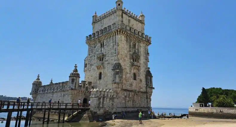 Uma torre de pedra histórica conhecida como Torre de Belém com telhado ameado, cercada por uma praia e água, sob um céu azul claro, com pessoas por perto. Representa roteiro em Portugal.