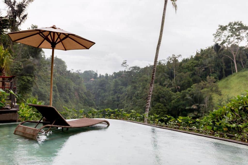 Imagem do Ulun Ubud Resort área à beira da piscina com espreguiçadeira e guarda-sol, com vista para uma floresta verdejante e colinas sob um céu nublado.