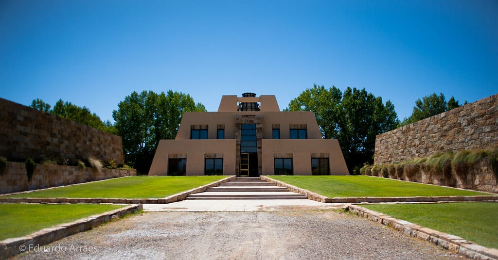 A Vinícola Catena Zapata em formato de pirâmide, com grandes janelas, é cercado por gramados verdes e muros de pedra sob um céu azul claro. Foto para ilustrar post sobre o que fazer em Mendoza.