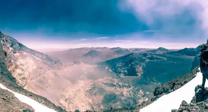 Nossa colunista está em uma cordilheira nevada, com vista para uma vasta paisagem rochosa com picos distantes sob um céu azul claro no vulcão Láscar no Deserto do Atacama.