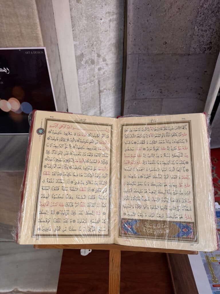 Um livro aberto com texto em árabe em um suporte de madeira, páginas cobertas com plástico protetor. Esse é um alcorão da Mesquita Süleymaniye.