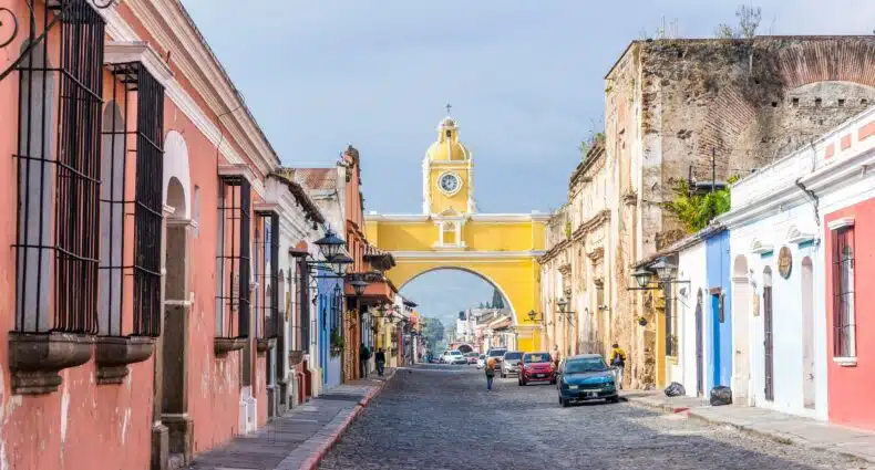Uma rua de paralelepípedos em Antígua ladeada por prédios coloridos leva a um arco amarelo com uma torre de relógio. Algumas pessoas e carros estão presentes. Representa o que fazer na Guatemala.