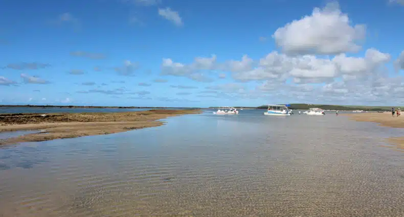 Praia Barra de São Miguel com águas rasas em primeiro plano e vários pequenos barcos flutuando sob um céu azul com nuvens dispersas. Em vários pontos é possível ver bancos de areia formados. A foto ilustra o post de o que fazer na Barra de São Miguel.