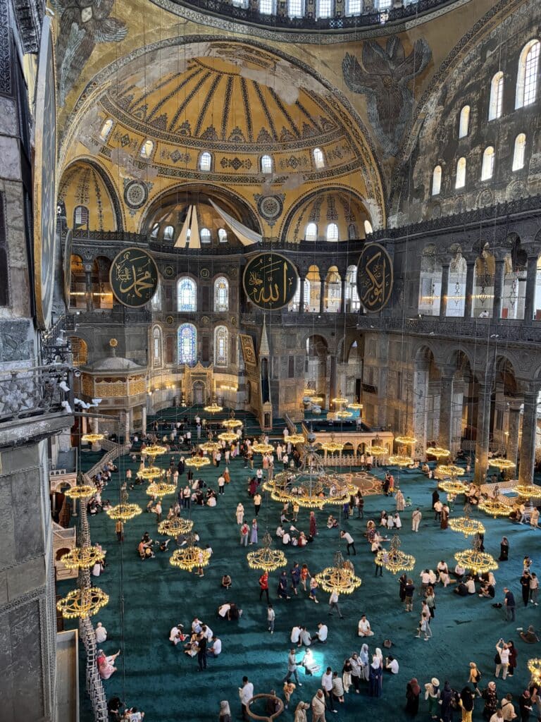 Interior de um grande edifício histórico com teto abobadado, designs ornamentados, lustres e pessoas reunidas em um tapete verde. Essa é a Basílica de Santa Sofia, uma das opções de o que fazer em Istambul.