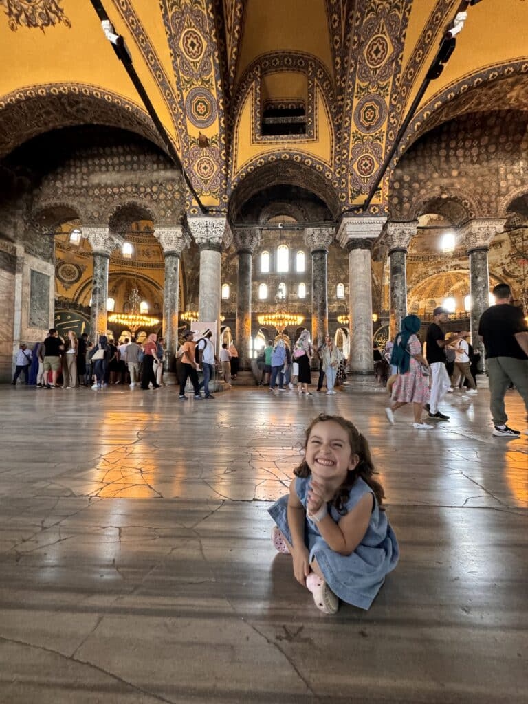 Uma menina está sentada no chão e sorri dentro da Basílica de Santa Sofia, um prédio grande e ricamente decorado, com tetos altos e uma multidão ao fundo.
