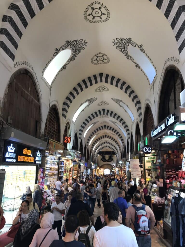 Um movimentado mercado interno com tetos altos e arqueados e designs ornamentados. As pessoas fazem compras entre várias lojas e barracas bem iluminadas no Bazar das Especiarias.