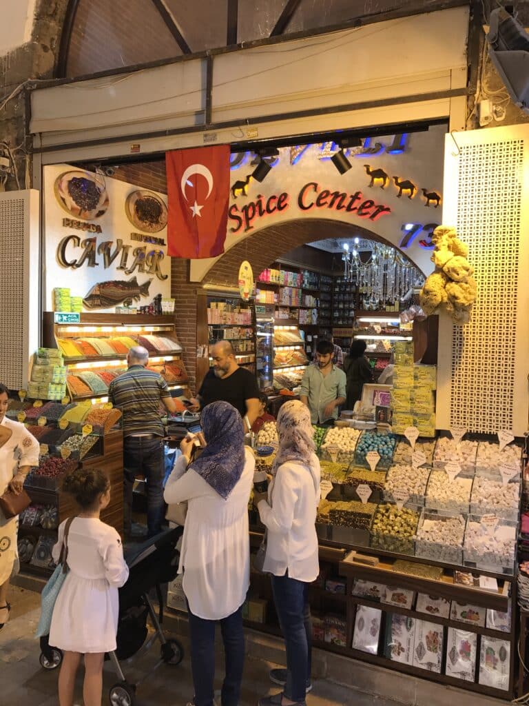 Pessoas compram em uma loja de especiarias e caviar com uma bandeira turca no Bazar das Especiarias, uma das opções de o que fazer em Istambul.. Especiarias e produtos são exibidos em prateleiras e balcões.
