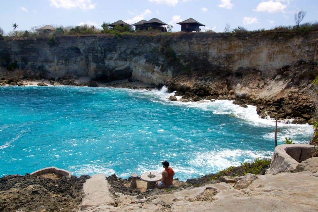 Pessoa sentada em uma rocha com vista para a Blue Lagoon, uma baía azul-turquesa, cercada por penhascos e ondas, com cabanas visíveis no topo dos penhascos.