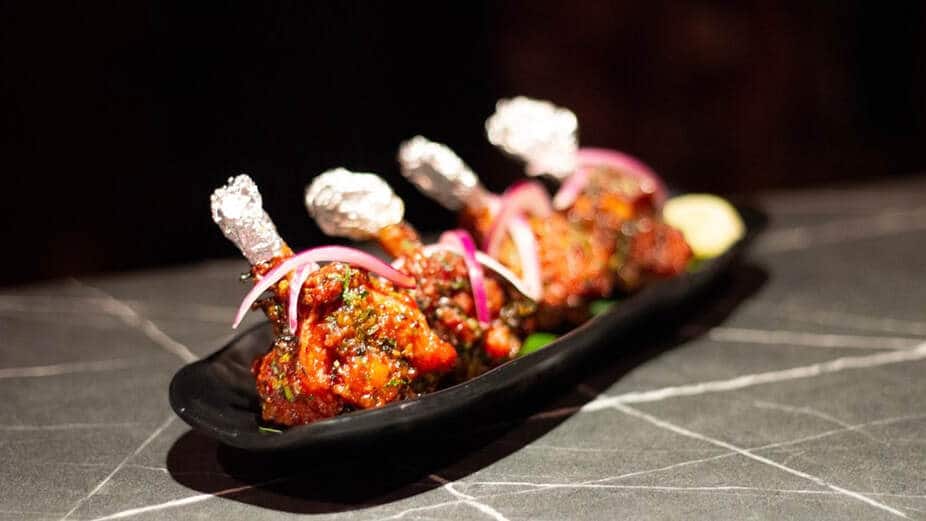 Close de um prato com três pirulitos de frango embrulhados em papel alumínio, guarnecidos com ervas e fatias de cebola em um prato preto.