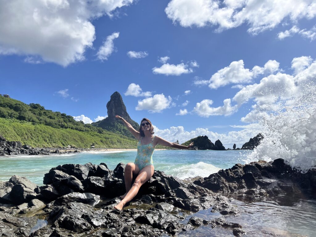 reveillon 2017 fernando de noronha foto: Virginia Falanghe Uma mulher de maiô sentada em pedras pretas perto do oceano, com ondas quebrando e uma grande colina rochosa ao fundo.