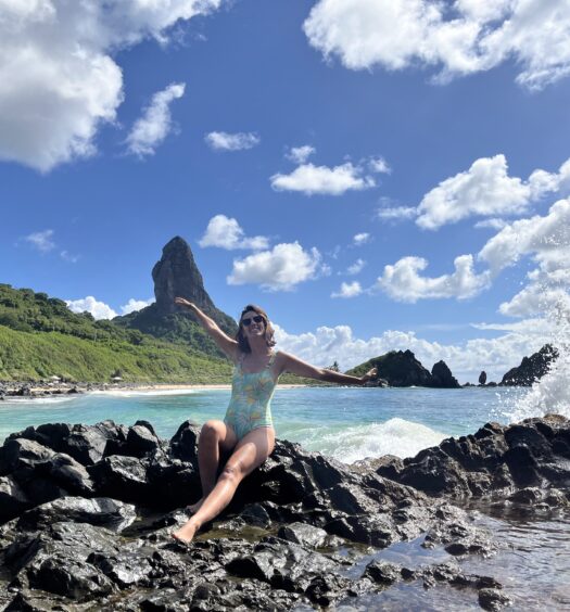 Uma mulher de maiô sentada em pedras pretas perto do oceano, com ondas quebrando e uma grande colina rochosa ao fundo.