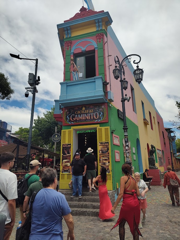 Um edifício colorido em uma rua de paralelepípedos com turistas ao redor; uma pessoa olha da sacada acima da entrada.