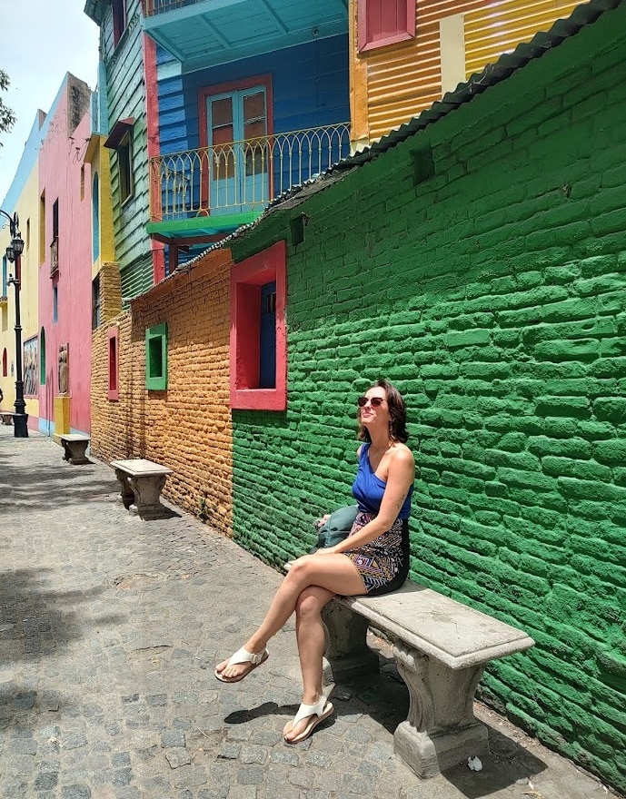 Pessoa sentada em um banco ao longo de uma rua colorida com edifícios vibrantes e multicoloridos ao fundo.