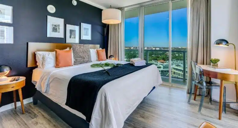 Um quarto de hotel moderno com uma cama grande, travesseiros, mesas de cabeceira, uma cadeira de escritório e uma grande janela com vista para a cidade. Representa hotéis bem localizados em Miami.