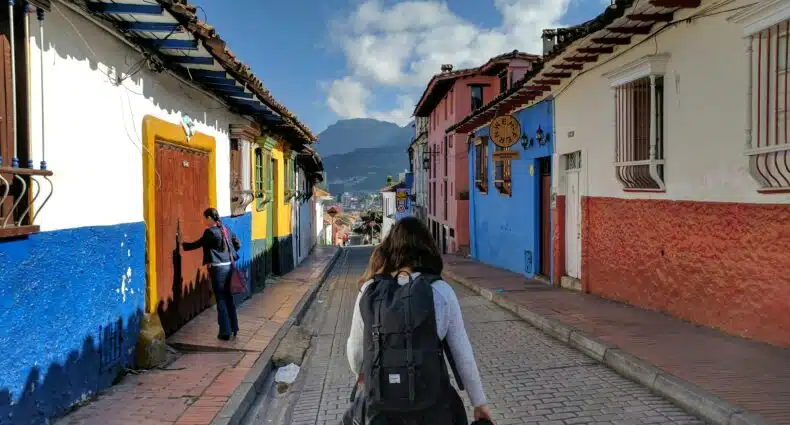 Uma pessoa com uma mochila caminha por uma rua colorida com prédios pintados, enquanto outra pessoa pinta uma porta. Montanhas são visíveis, em Bogotá. Representa o que fazer em Bogotá.