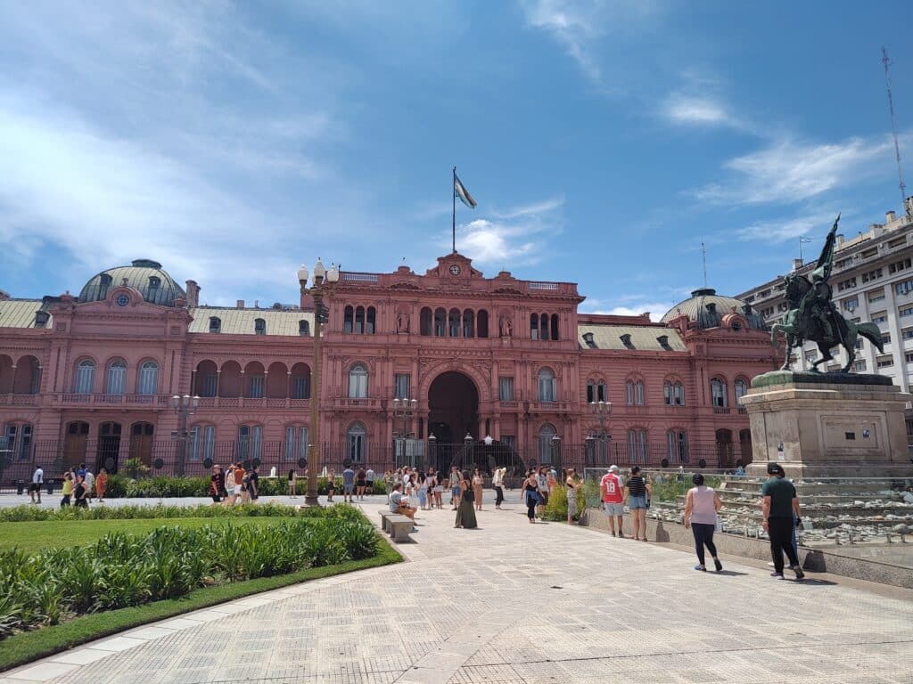 Um grande edifício rosa com uma bandeira no topo sob um céu azul. Pessoas andam perto de uma estátua em primeiro plano.