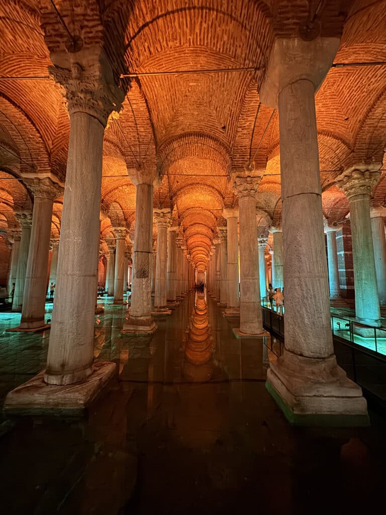 A imagem mostra um salão subterrâneo com colunas e água refletida na Cisterna da Basílica, iluminada por luzes quentes e frias. Essa é uma das opções de o que fazer em Istambul.