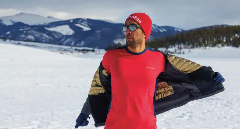 Uma pessoa está em uma paisagem nevada vestindo uma camisa vermelha, gorro vermelho e óculos de sol, com montanhas ao fundo, para representar o post de Columbia outlet