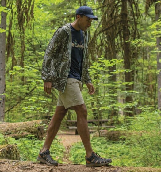 Um homem de boné caminha sobre um tronco em uma floresta verdejante e exuberante. Ele veste uma jaqueta corta vento Columbia, shorts e tênis, cercado por árvores e plantas altas.