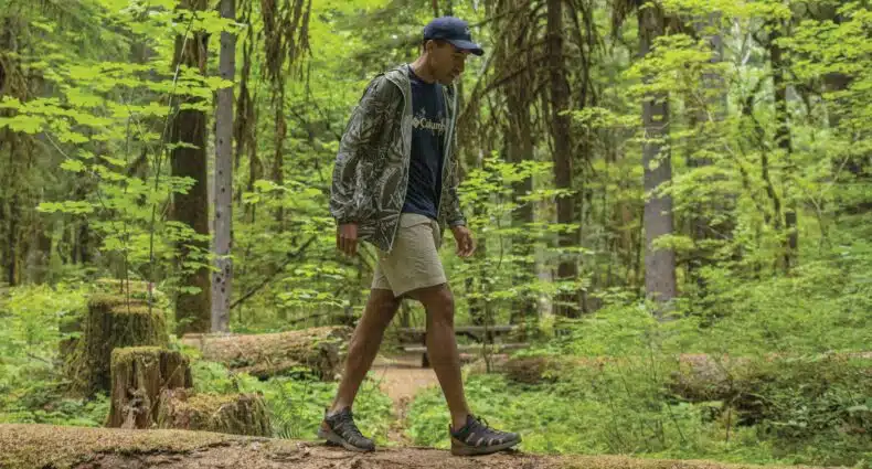 Um homem de boné caminha sobre um tronco em uma floresta verdejante e exuberante. Ele veste uma jaqueta corta vento Columbia, shorts e tênis, cercado por árvores e plantas altas.