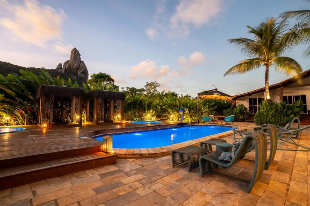 Um quintal com piscina, espreguiçadeiras, um deck de madeira e uma palmeira no Dolphin Hotel. Colina rochosa e céu nublado ao fundo.