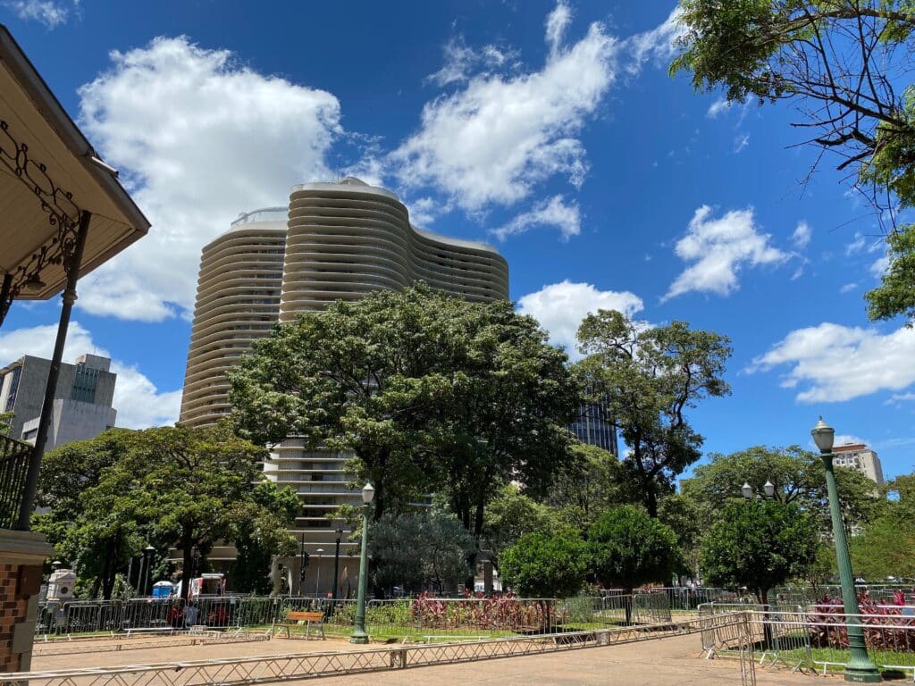 Prédio alto com fachada ondulada atrás de árvores, sob um céu azul com nuvens. Um parque e um pavilhão estão em primeiro plano.