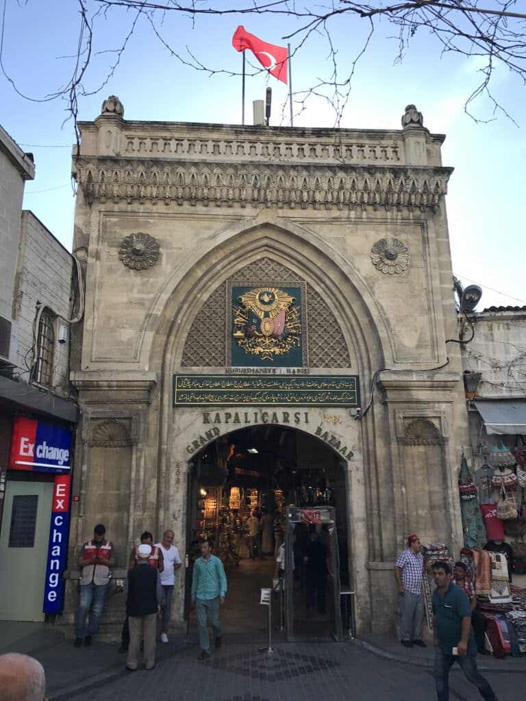 A entrada do Grande Bazar em Istambul, com um arco ornamentado e a bandeira turca no topo. Pessoas estão caminhando por perto. Essa é uma das opções de o que fazer em Istambul.