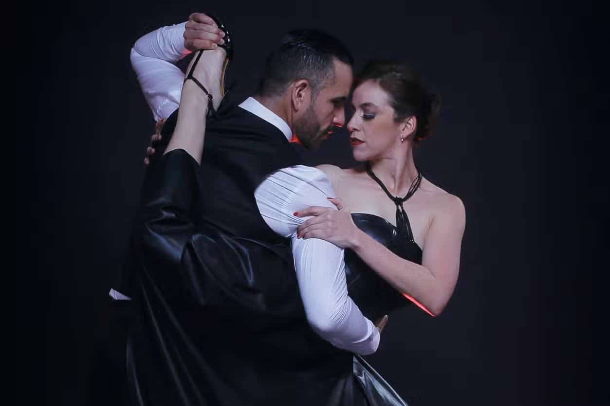 Um homem e uma mulher dançam juntos em trajes formais, com a mulher executando um levantamento de perna alto contra um fundo escuro.