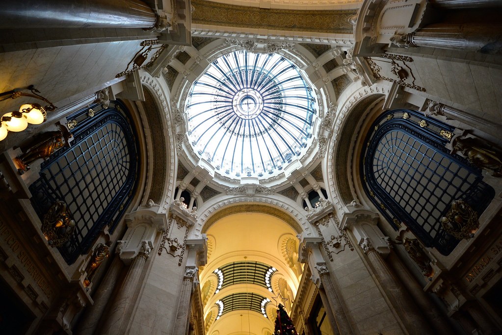 Vista interna da Galeria Guemes, um dos pontos turísticos de Buenos Aires, de um grande edifício com teto de vidro abobadado e arquitetura ornamentada com grandes arcos decorativos.