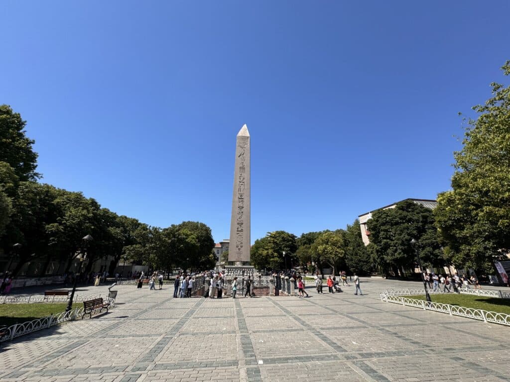 Um alto obelisco fica em uma praça pavimentada cercada por pessoas, com árvores e um céu azul claro ao fundo. Esse é o Obelisco de Teodósio na Praça Sultanahmet, uma das opções de o que fazer em Istambul.