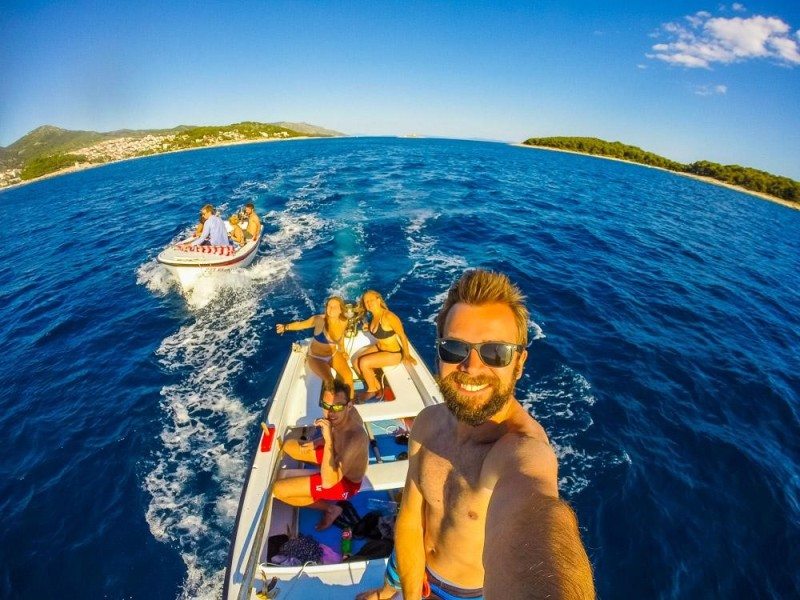 Pessoas sorrindo em um barco no oceano, em direção às Ilhas Paklene na Croácia, com uma pessoa tirando uma selfie. Outro barco com pessoas está ao fundo.