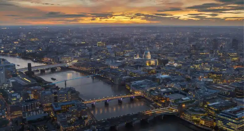 Vista aérea de Londres ao anoitecer, com um rio correndo por ela, várias pontes e edifícios iluminados sob um céu nublado. A imagem ilustra o post sobre roteiro para Londres.