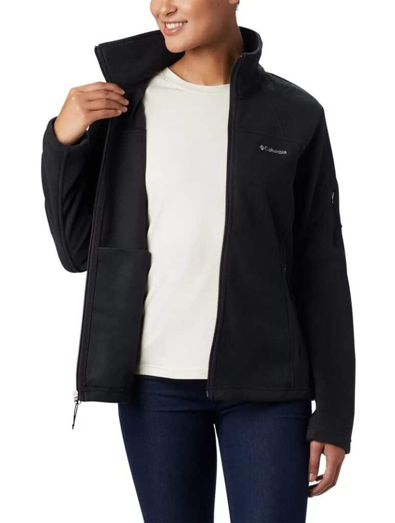 Uma pessoa vestindo uma camisa branca, jaqueta escura e jeans azul, está de pé com uma mão ajustando a gola da jaqueta. Essa é a Jaqueta Fleece Columbia Feminina Fast Trek II.