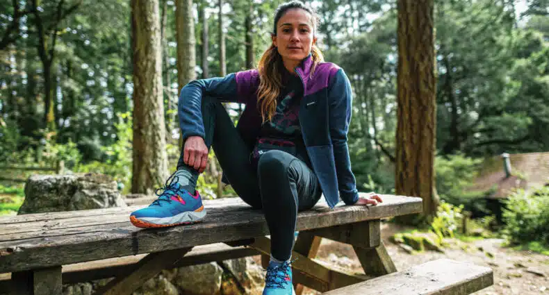 Uma mulher está sentada em uma mesa de piquenique de madeira em uma floresta, vestindo uma jaqueta Fleece Columbia roxa e azul, leggings e tênis de trilha coloridos.