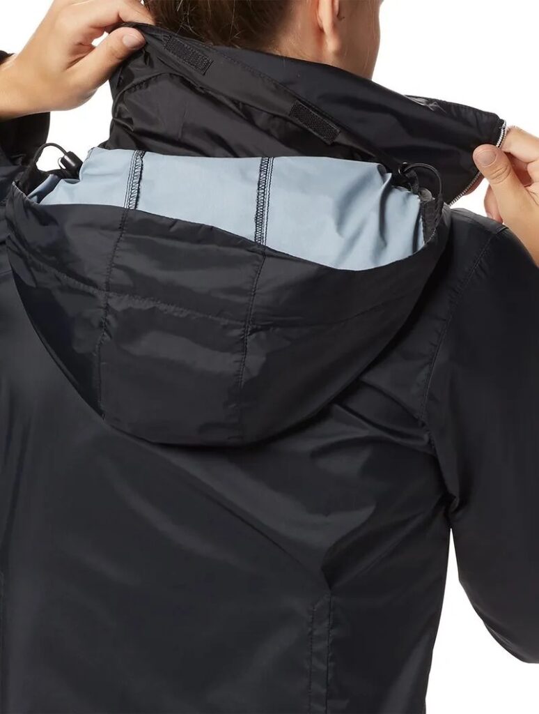 Uma pessoa ajusta o capuz de uma jaqueta preta com forro azul, vista de trás, contra um fundo branco. Essa é a Jaqueta Longa Columbia Feminina Switchback III.