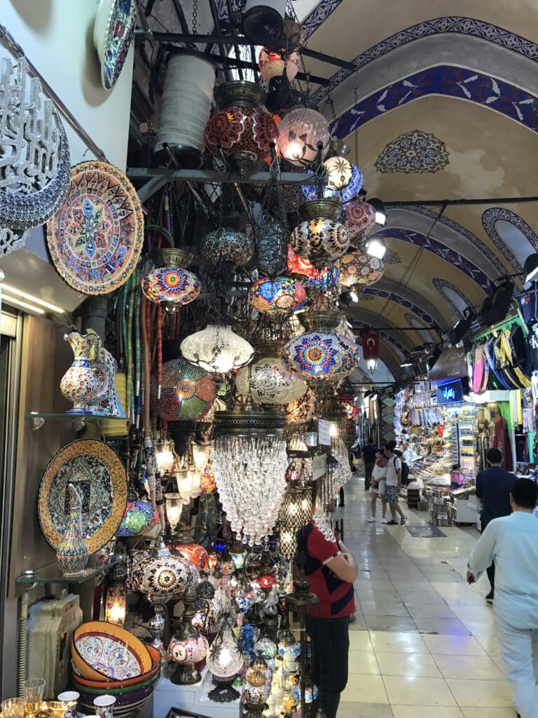 Uma barraca do Grande Bazar vibrante exibe luminárias de mosaico coloridas e pratos decorativos sob um teto abobadado com detalhes em azulejos.