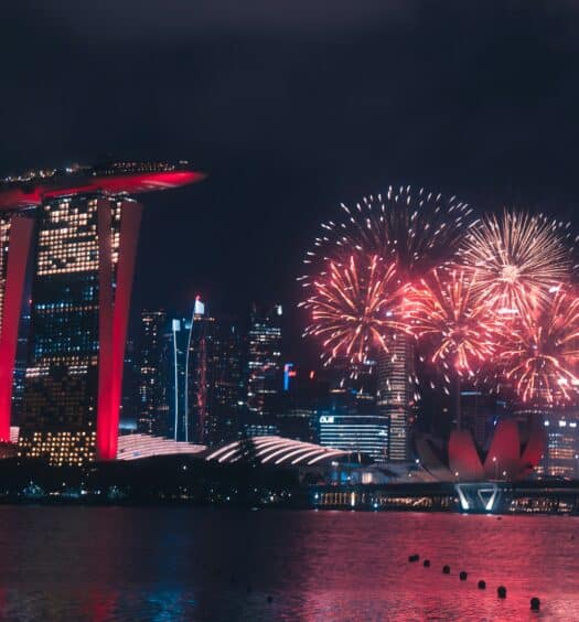 Fogos de artifício coloridos iluminam o céu noturno sobre Marina Bay Sands e prédios da cidade em Singapura, refletidos na água.